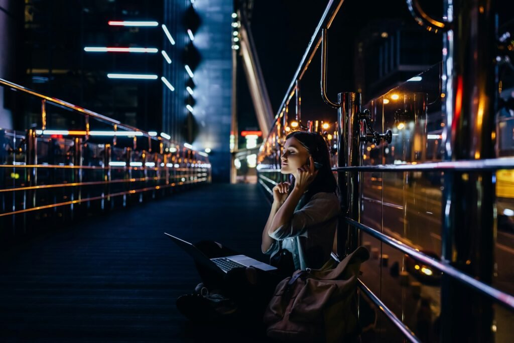 side view of woman listening music in headphones with laptop on knees on city street at night